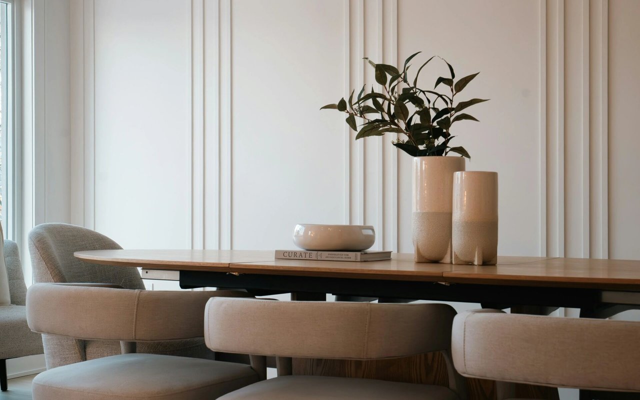 Dining room with a wooden table, upholstered chairs, and leafy branches in tall ceramic vases.