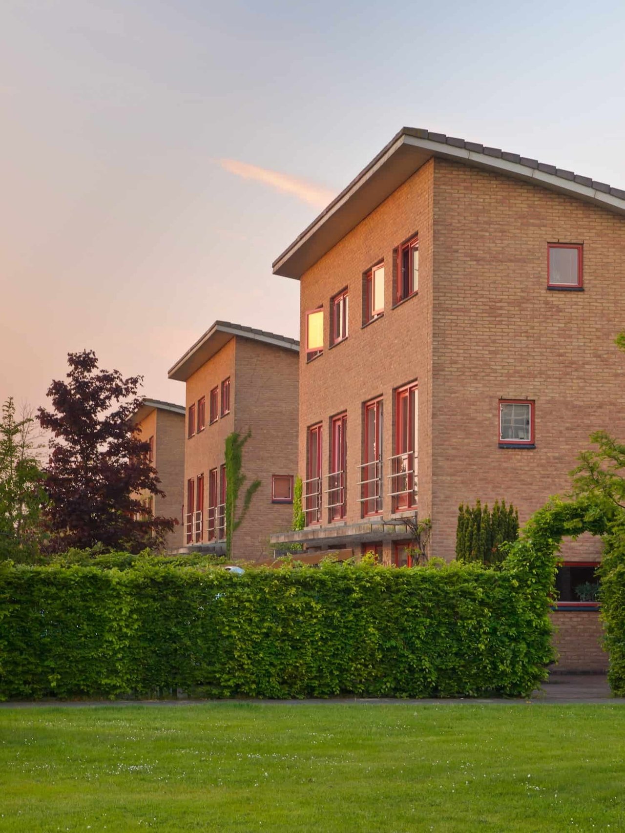 Brick residential buildings with red-trimmed windows rise behind a hedge and lawn in warm evening light.