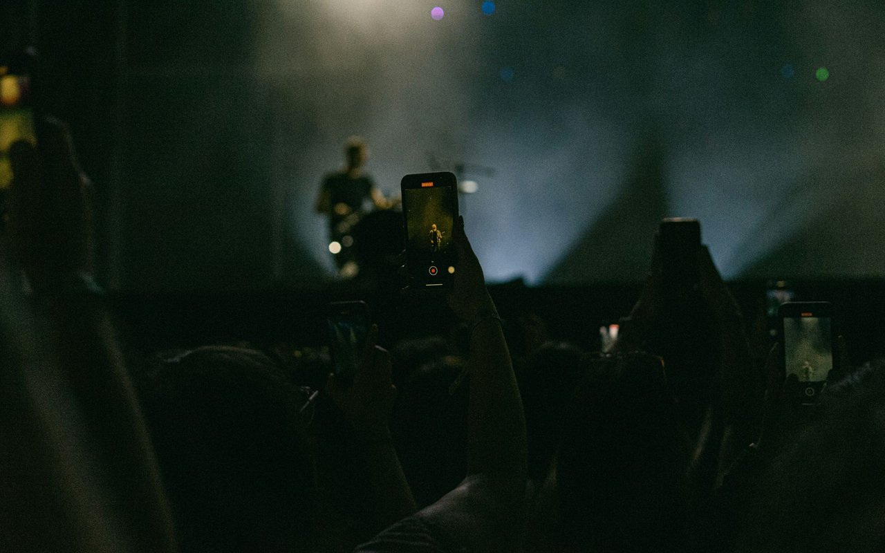 Concertgoers film a performer onstage with their phones in a dark crowd.