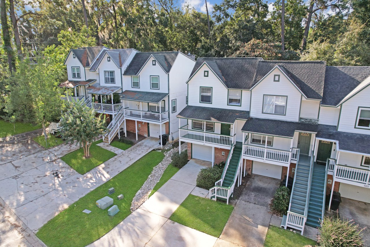 Aerial view of the townhome at 529 Collinsford Road, Tallahassee, Florida, showing a row of attached two-story homes with white siding, green trim, and covered front porches. Each unit has a one-car garage and a private staircase leading to the main entrance. The property is surrounded by mature trees and well-maintained landscaping.
