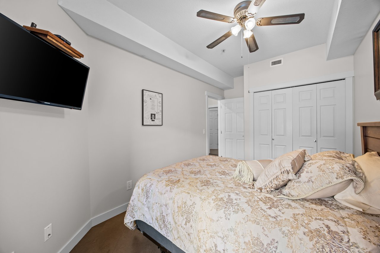 Furnished bedroom with a floral-quilted bed, wall-mounted television, and white closet doors.