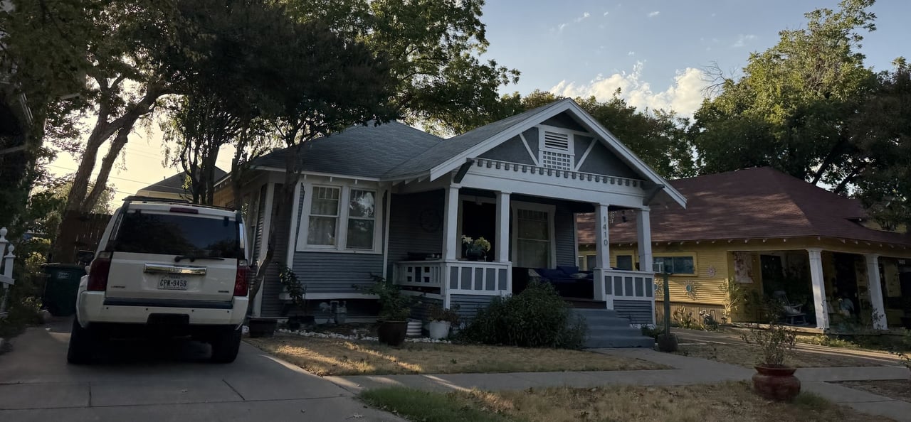 Gray bungalow with a white-columned front porch beneath mature trees.