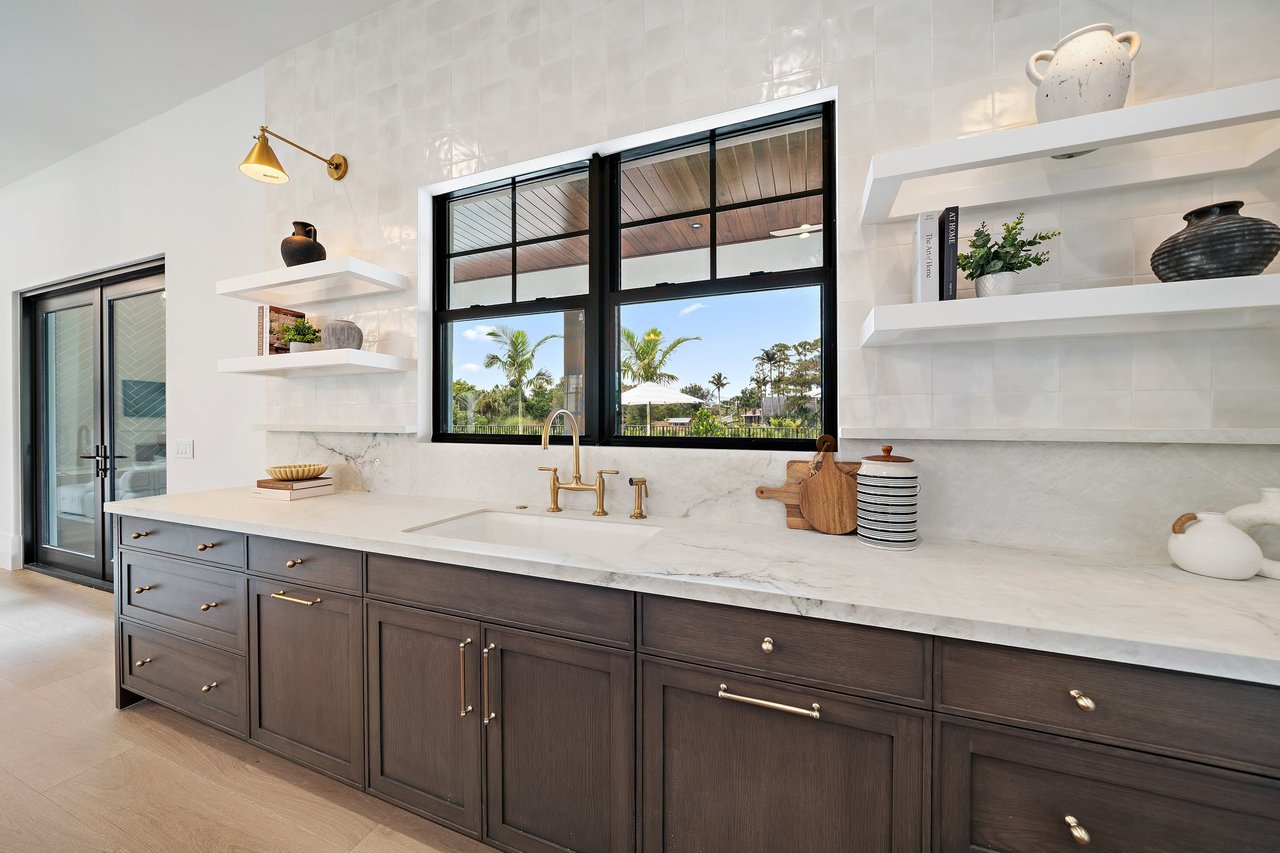 Modern kitchen with dark wood cabinets, brass fixtures, and a large window overlooking palm trees.