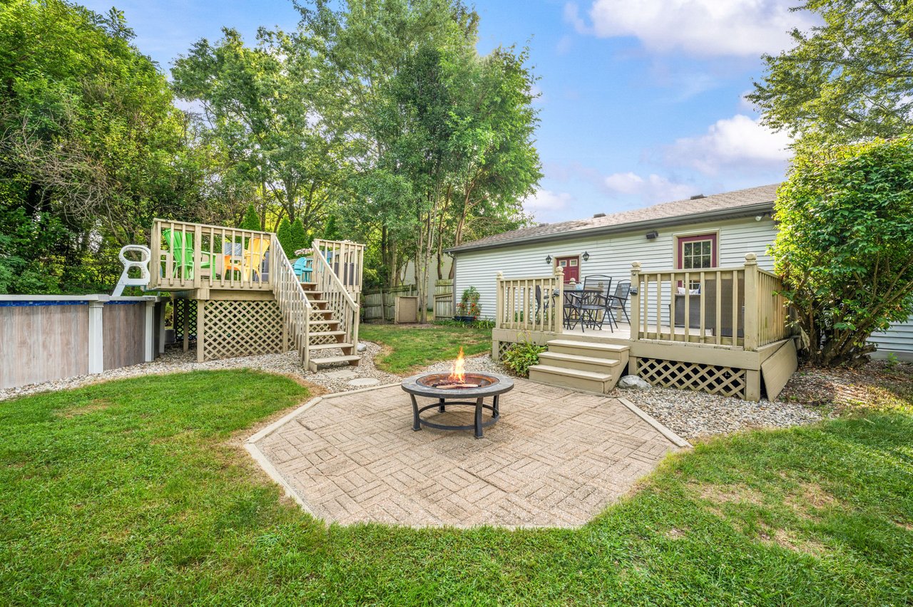 Backyard with a raised deck, above-ground pool, paved fire-pit patio, and mature trees.