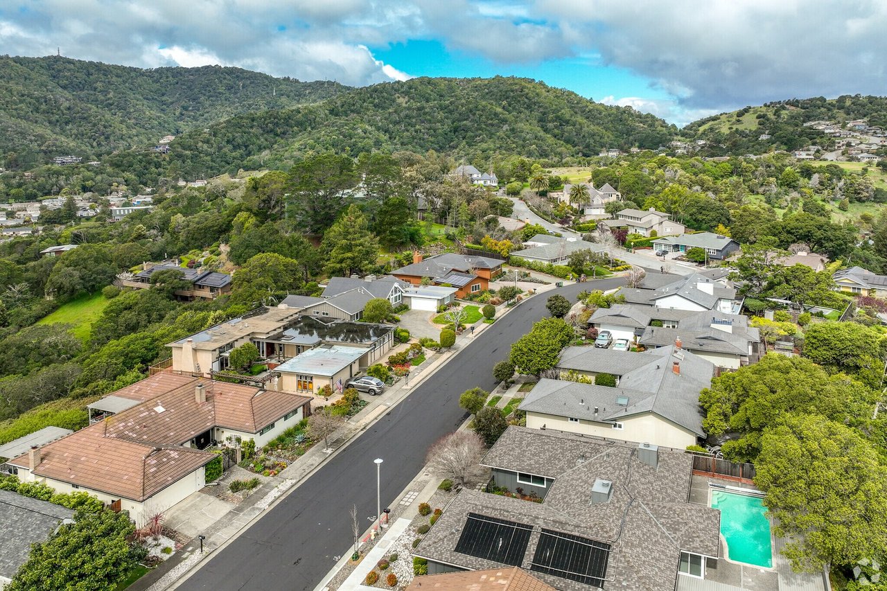 Aerial view of a residential neighborhood with a curving street, backyard pool, and wooded hills.