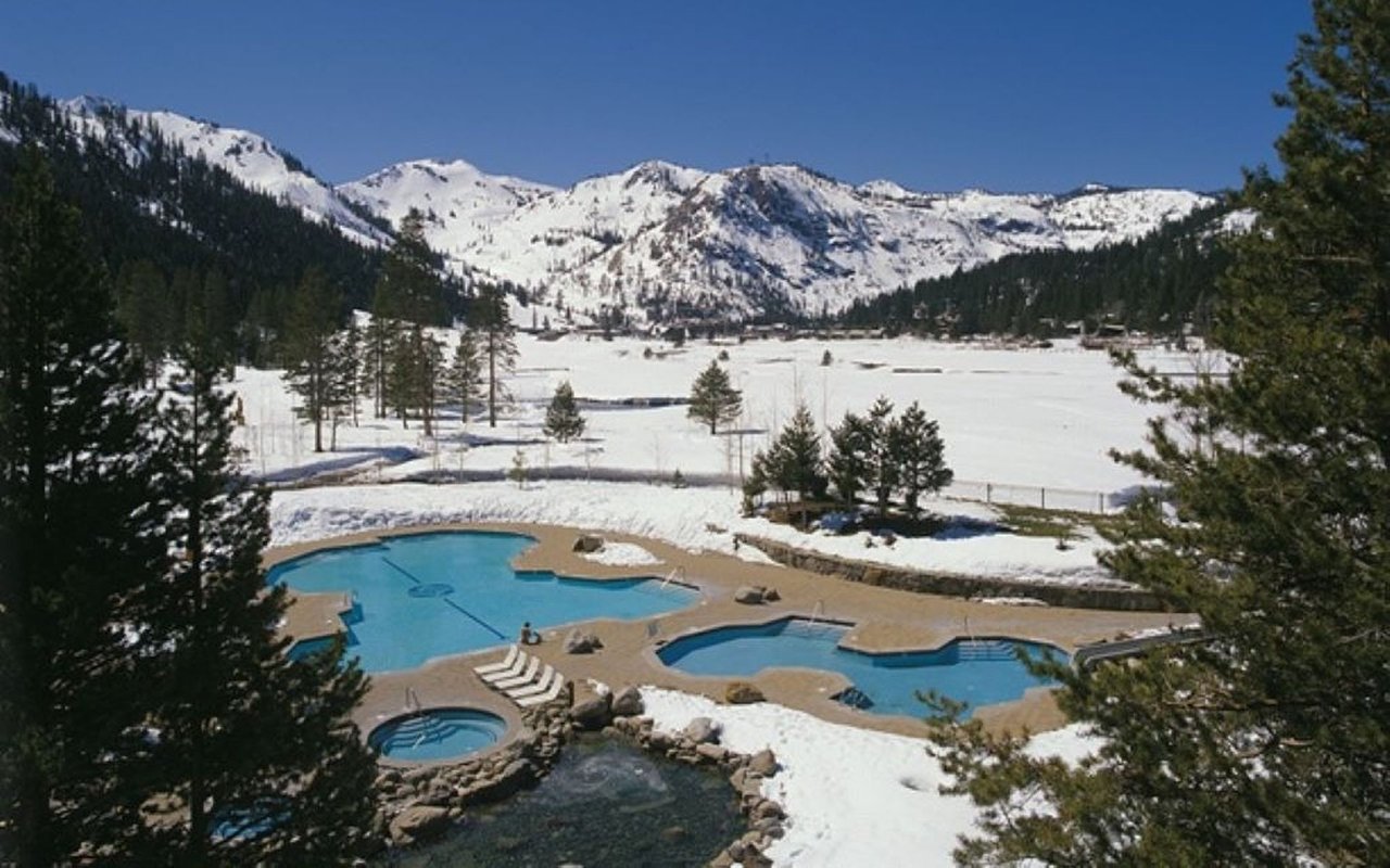 Resort pools and hot tub beside a snowy field, with evergreen trees and mountain peaks beyond.