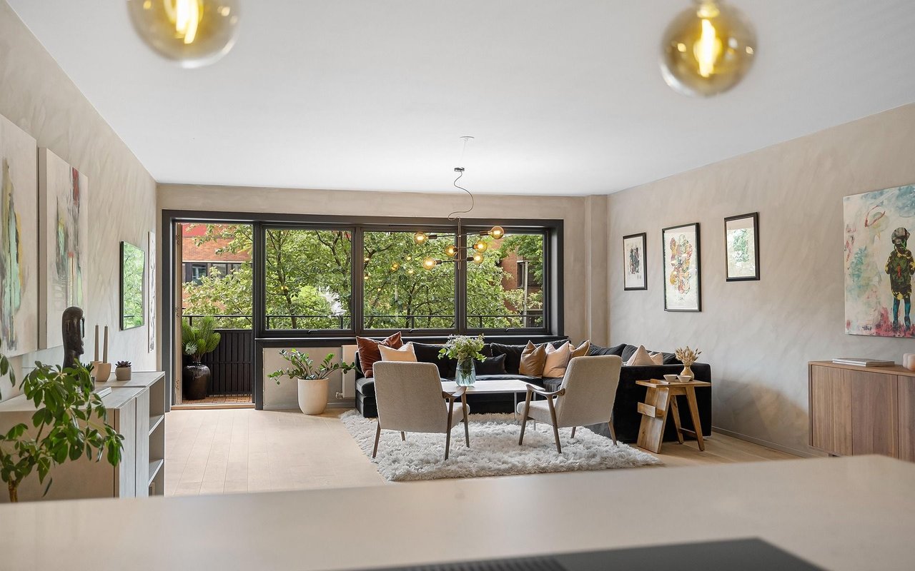 Living room with a black sofa, beige armchairs, colorful artwork, and large windows overlooking trees.