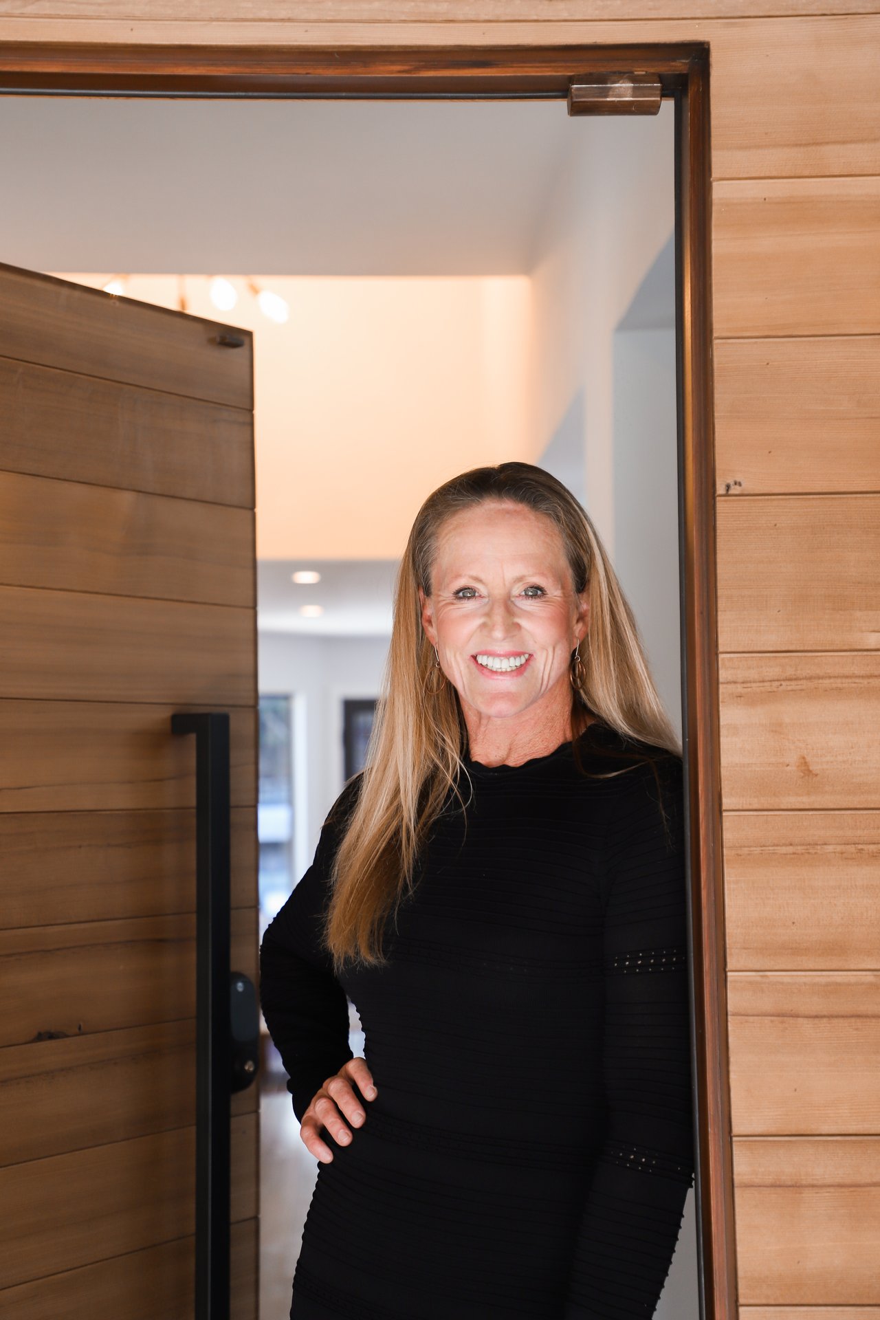 Smiling woman with long blonde hair in a black top, standing in a wooden doorway.