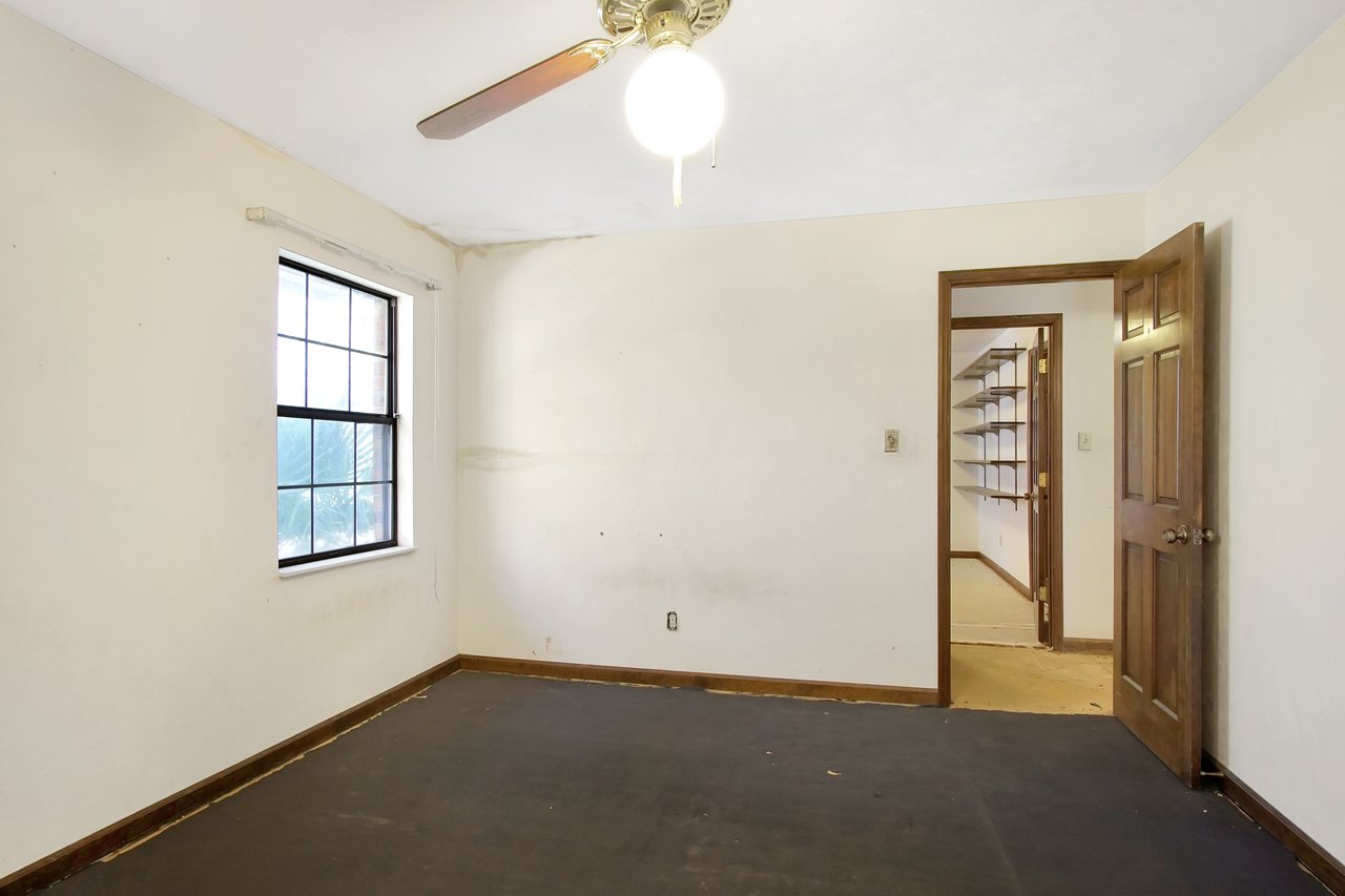 Empty room with white walls and a dark floor, featuring a window with a grid pattern and an open wooden door leading to a hallway with shelves. A ceiling fan with a light is on, creating a neutral, calm atmosphere.