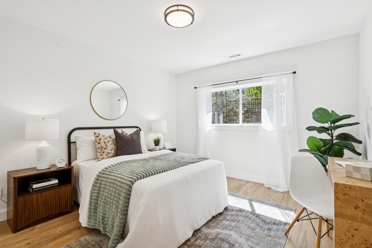 Staged bedroom with a white bed, sage-green throw, round mirror, and sunlit window.