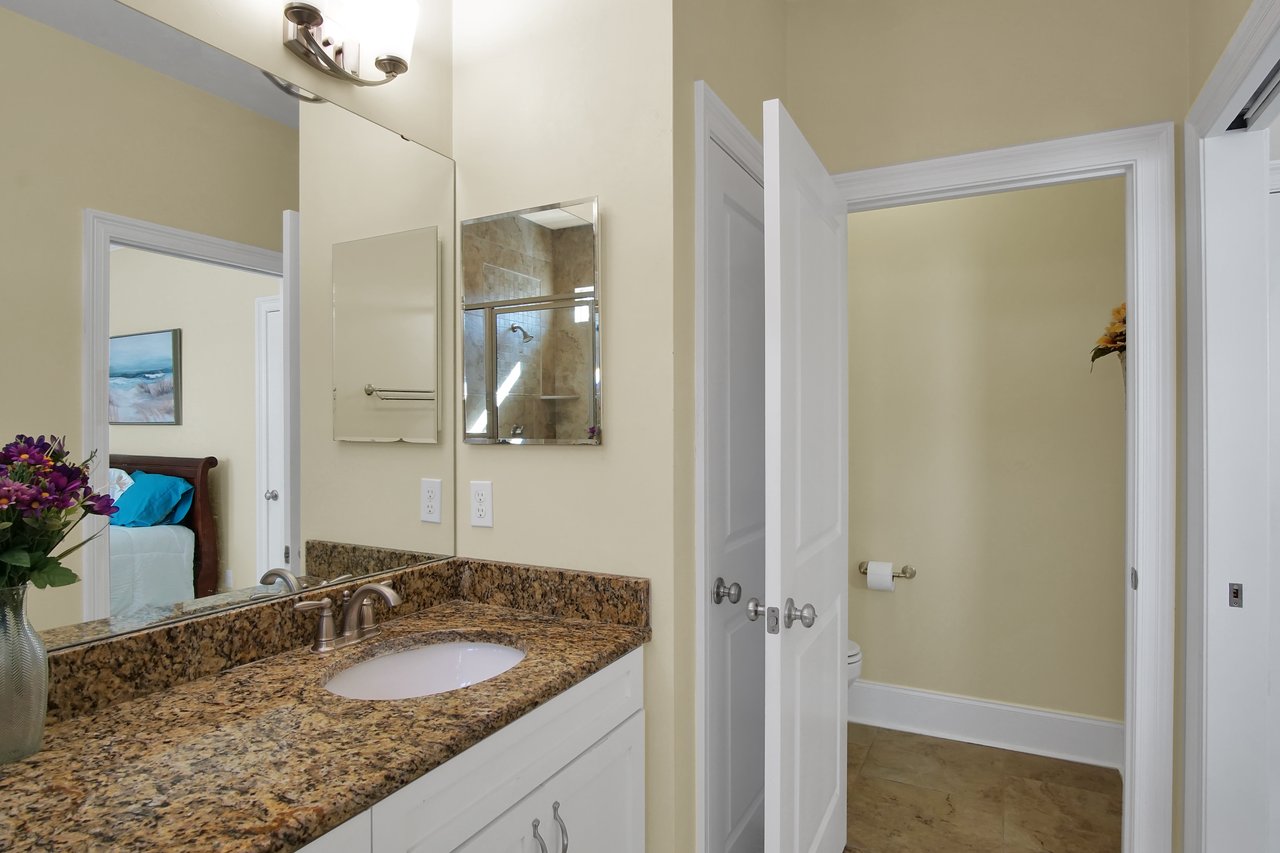 Bathroom with a granite countertop and sink on the left, double doors leading to a toilet on the right. A vase with purple flowers adds color.