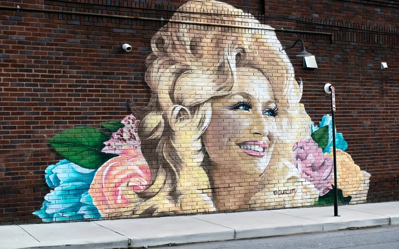 A mural of a smiling woman with blue eyes surrounded by colorful flowers, and is painted on a red brick wall.