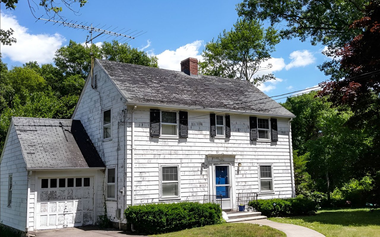 Weathered two-story white house with peeling siding, dark shutters, and an attached garage.