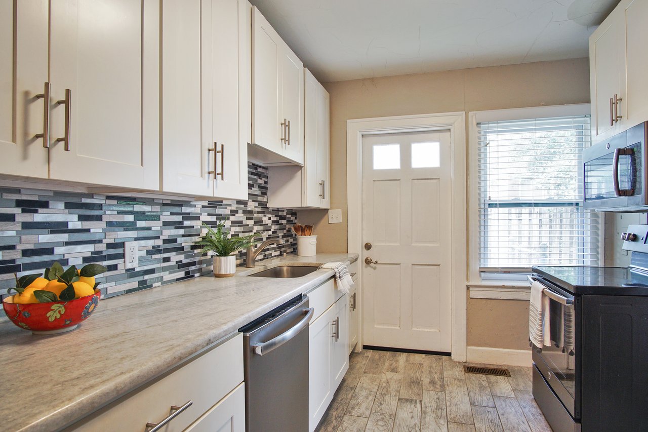 Bright kitchen with white cabinets, gray stone countertops, and a mosaic tile backsplash. A bowl of lemons and potted plant add color. Airy, modern feel.