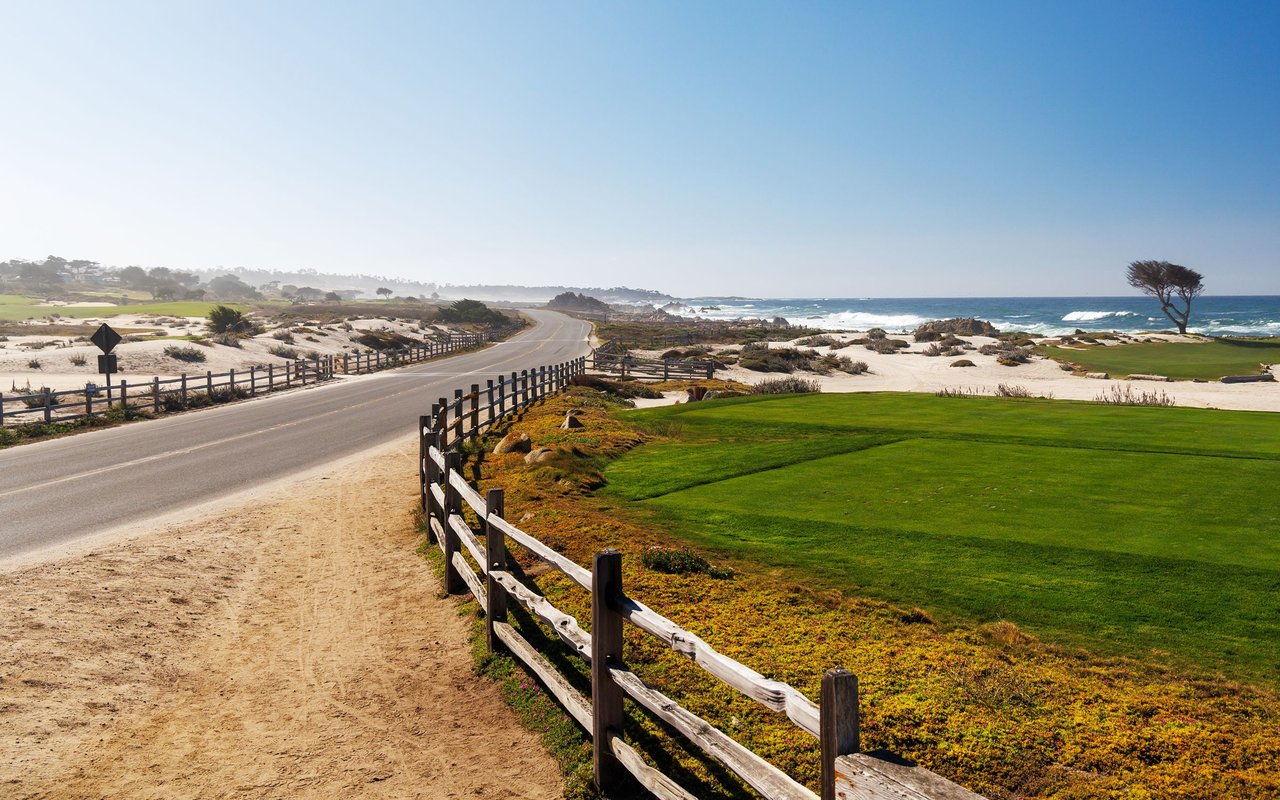 Coastal golf course beside a curving road, with sandy dunes, wooden fences, and ocean waves.