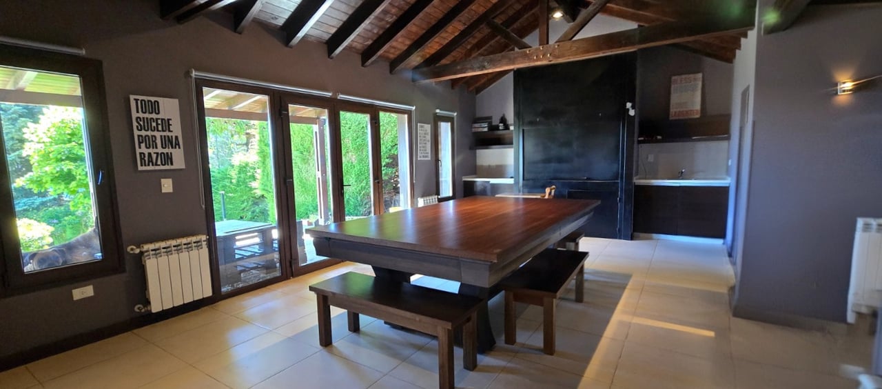Dining area with a large wooden table, exposed beams, glass doors, and a dark kitchen.