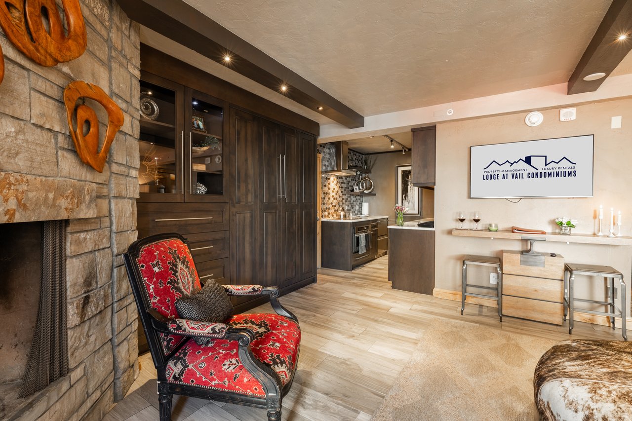 Condominium living area with a stone fireplace, red patterned armchair, and dark wood cabinetry.