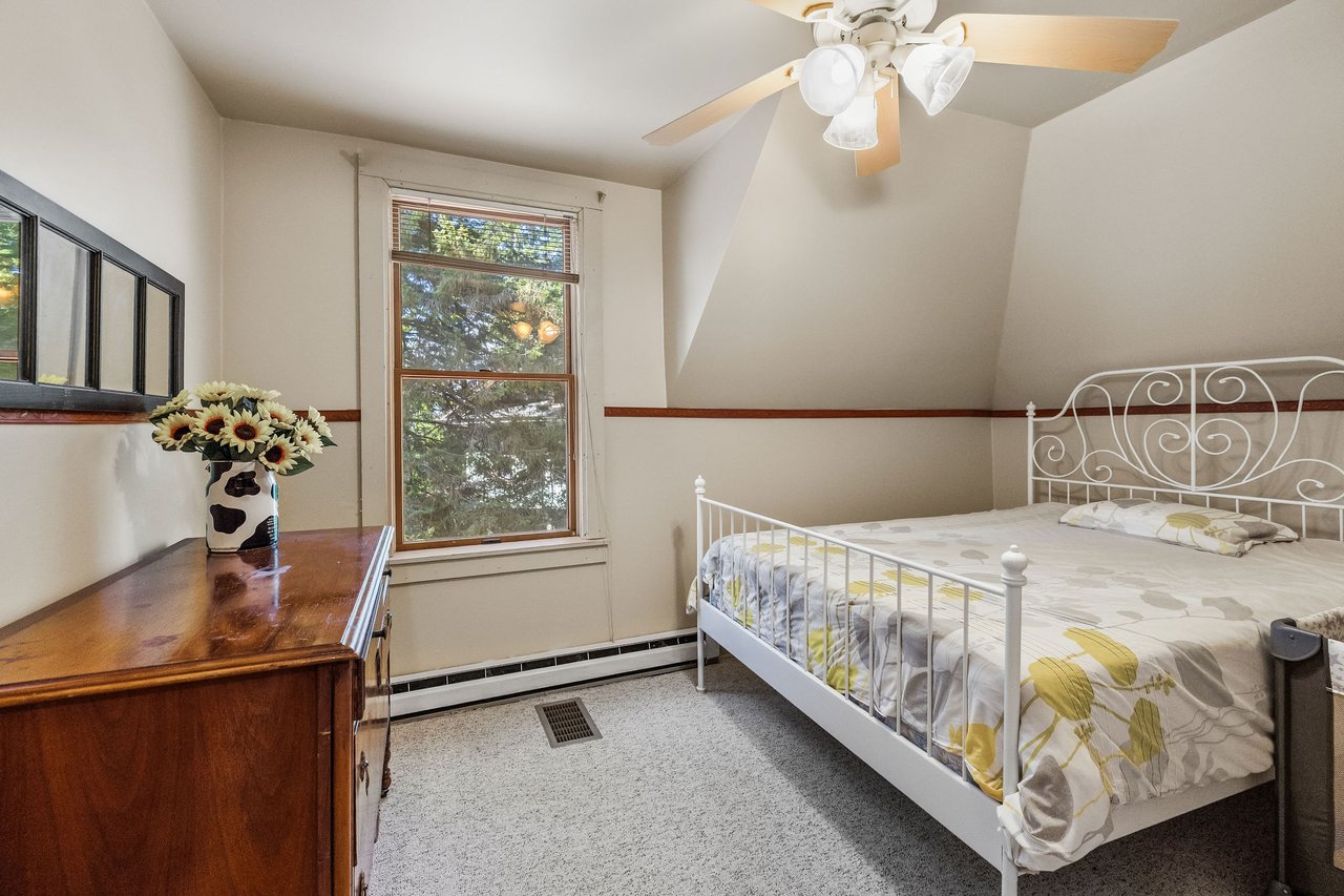 Bedroom with a white metal bed, wooden dresser topped with sunflowers, and a window overlooking trees.