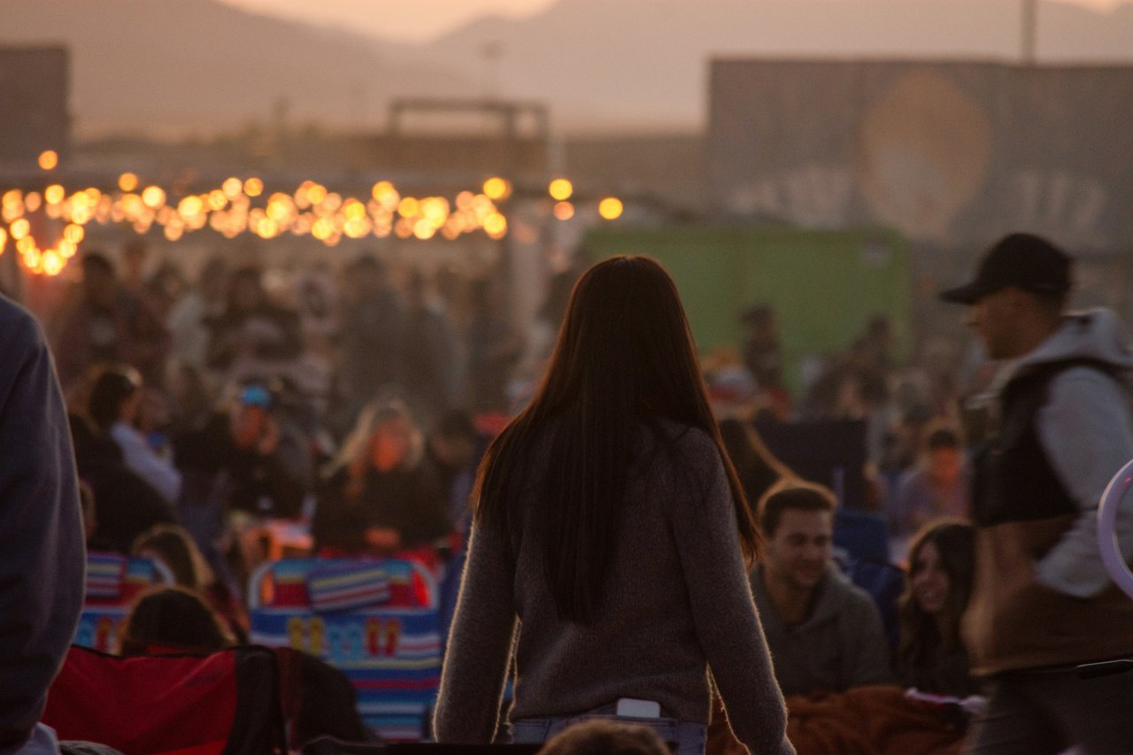 A woman standing in a crowd at an outdoor summer event at dusk with warm string lights in the background