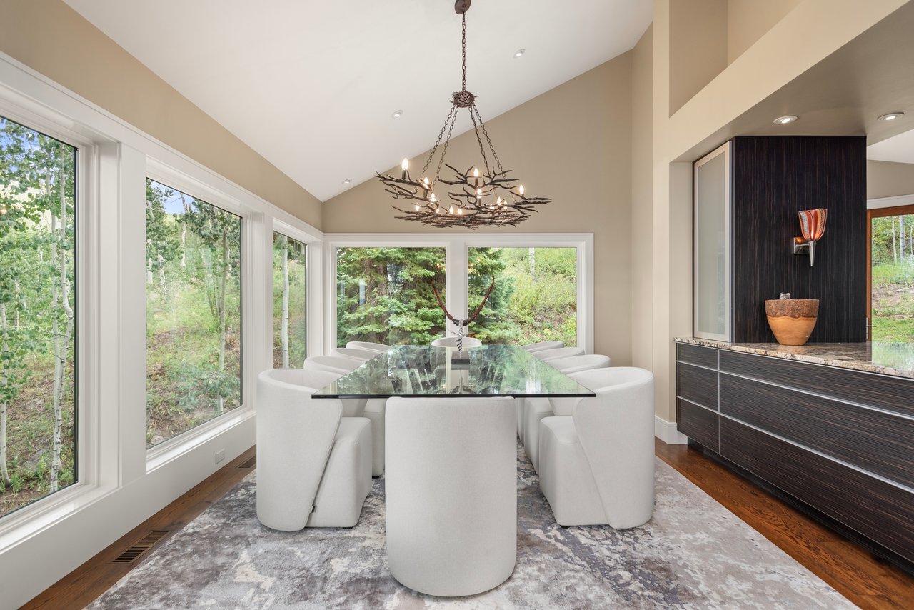 Dining room with a glass-top table, white chairs, branch chandelier, and windows overlooking the woods.