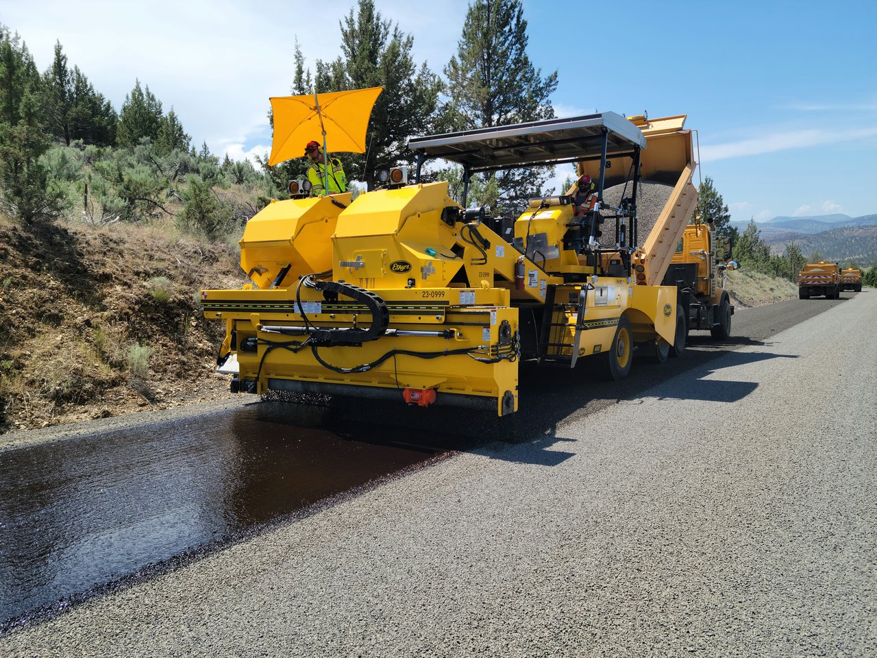 Yellow road-treatment truck spreading a dark coating across a rural road, with workers operating the machinery.