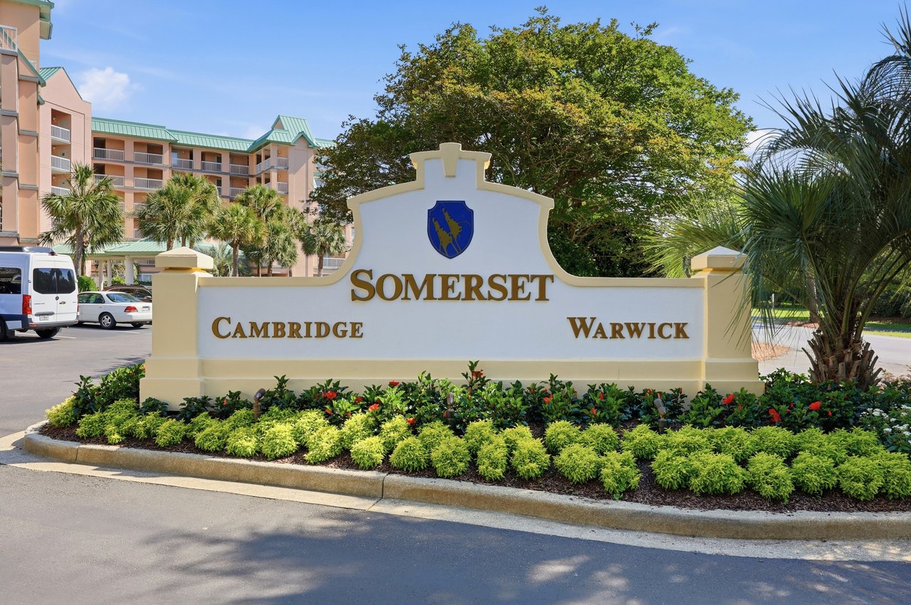 Somerset entrance sign naming Cambridge and Warwick, with palm trees and multistory buildings behind it.