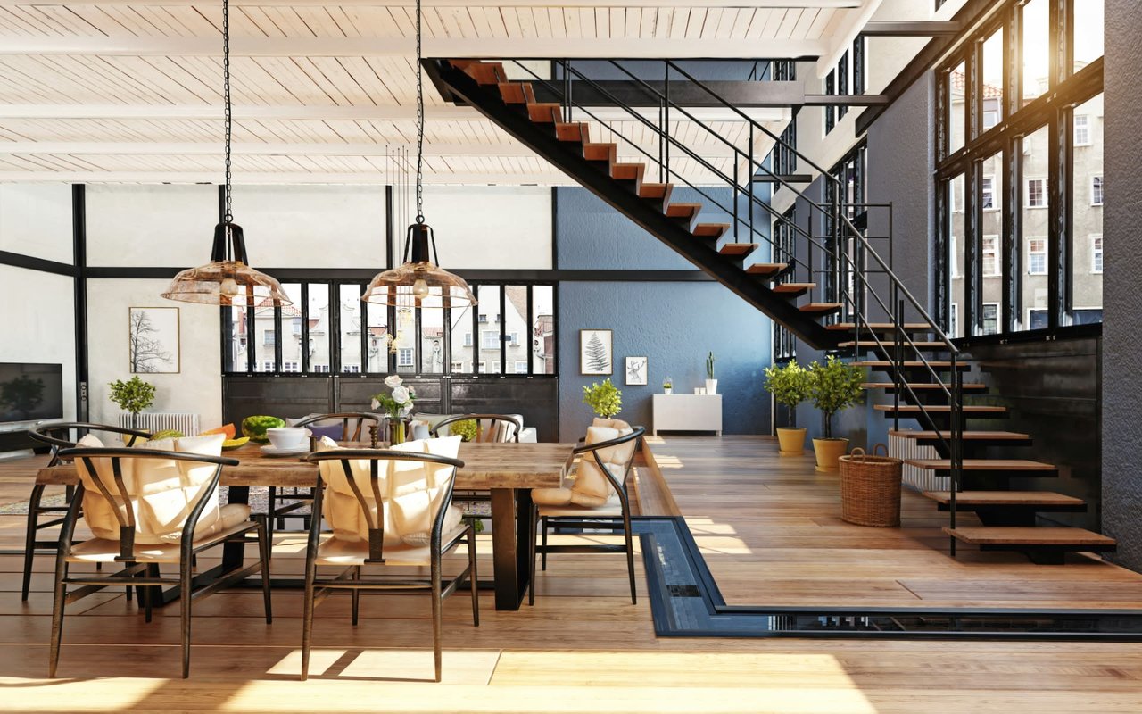 A dining area with a wooden table and chairs in the foreground, with a staircase leading up to the second floor behind it.