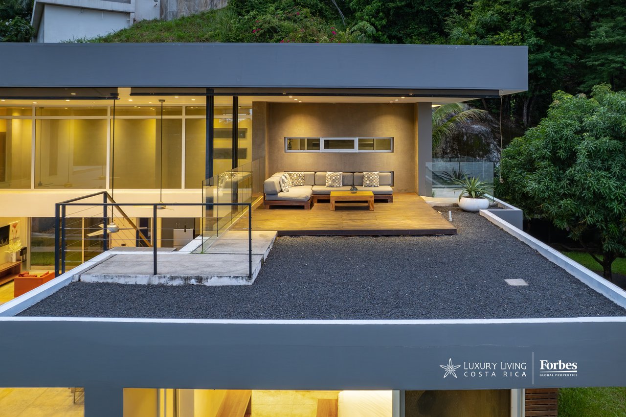 Modern glass-fronted home with a furnished terrace, warmly lit among tropical greenery.