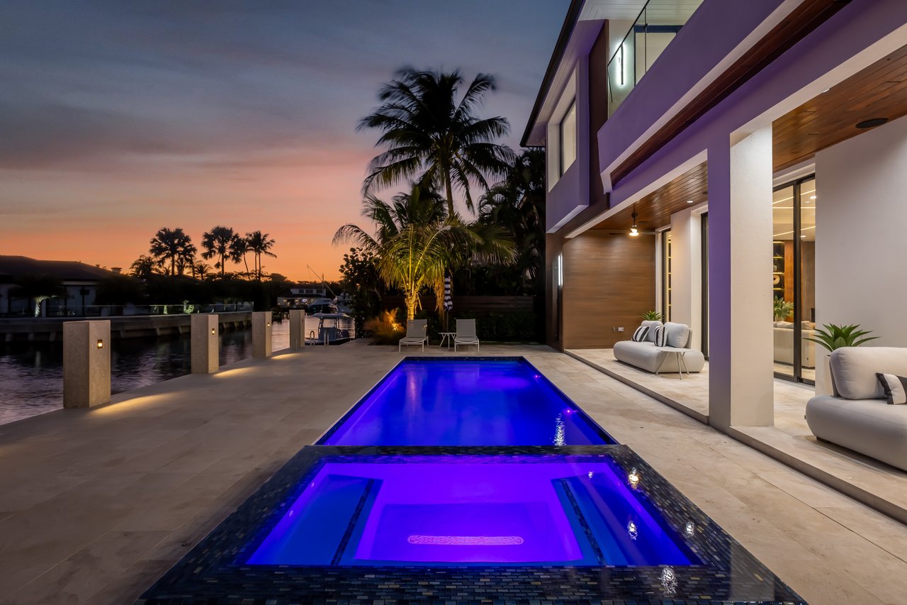 Modern waterfront home with a blue-lit pool and spa beside a canal at sunset.