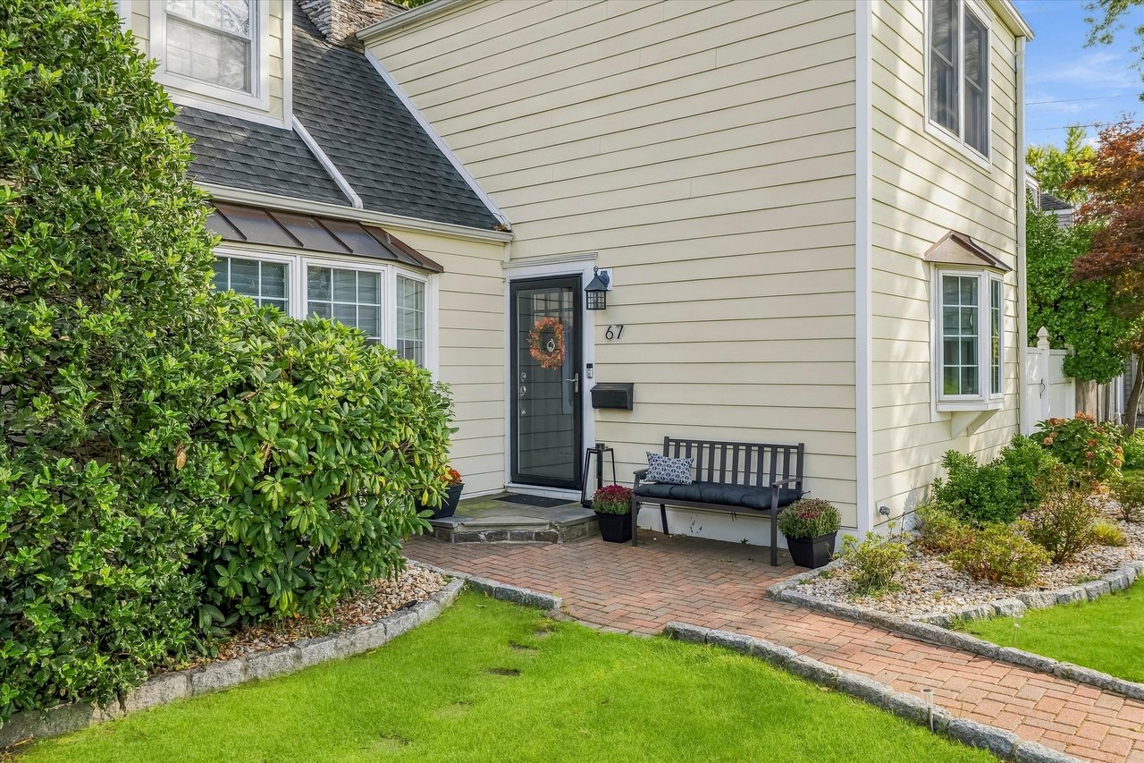 House entrance marked 67, with a brick walkway, garden beds, and a bench beside the door.