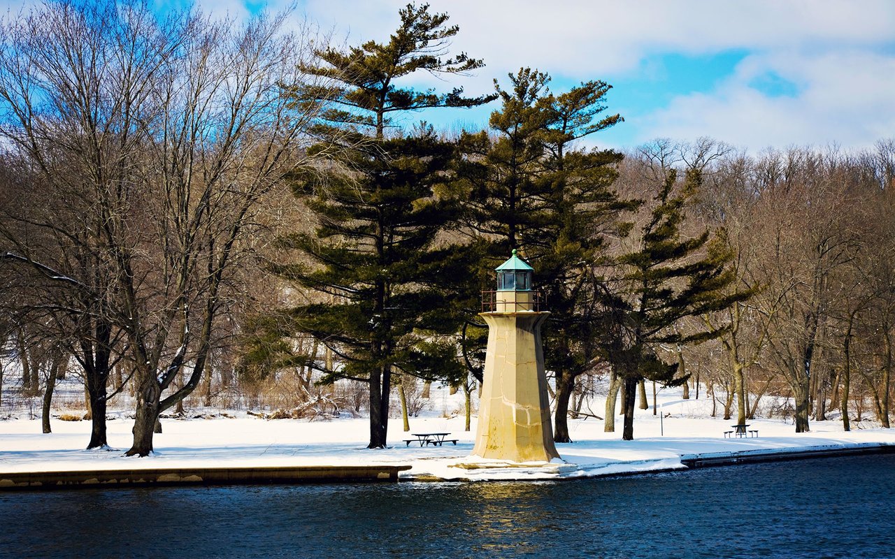 Small lighthouse beside a river in a snowy park, surrounded by bare trees and pines.