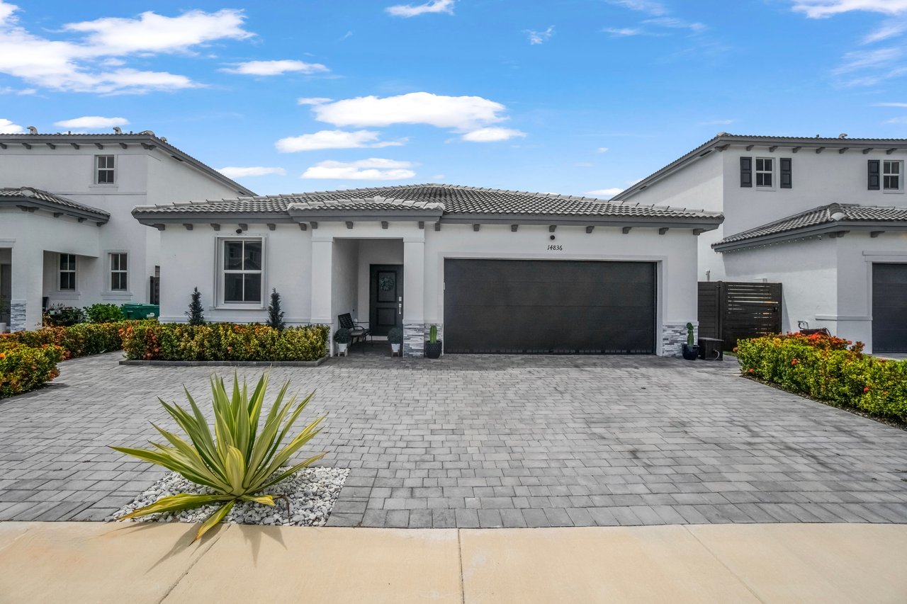 White stucco home with a gray tile roof, dark double garage door, and broad paved driveway.