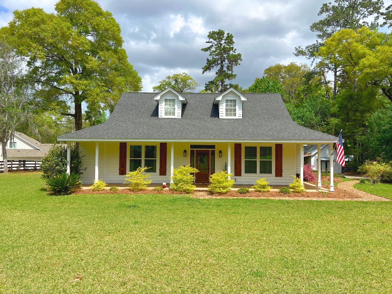 Charming white house with a gray roof, front porch, and American flag. Surrounded by vibrant greenery and trees under a partly cloudy sky.