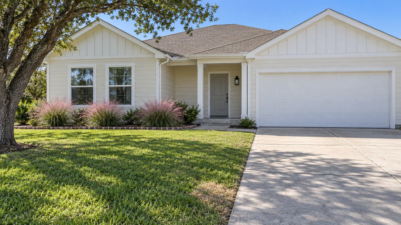 Cream-sided single-story home with taupe gables, a shallow porch, white garage door, broad green lawn and an oak tree in clear afternoon light.