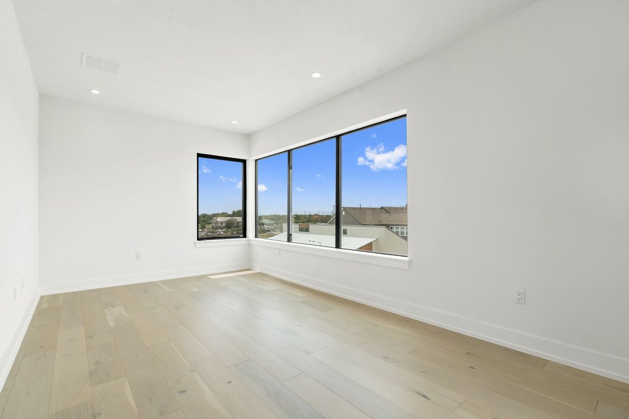 Empty room with pale wood floors and large corner windows overlooking nearby homes.