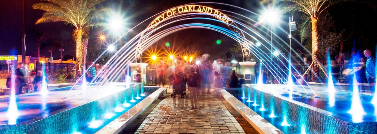 City of Oakland Park arch and fountain plaza at night with lights and palm trees.