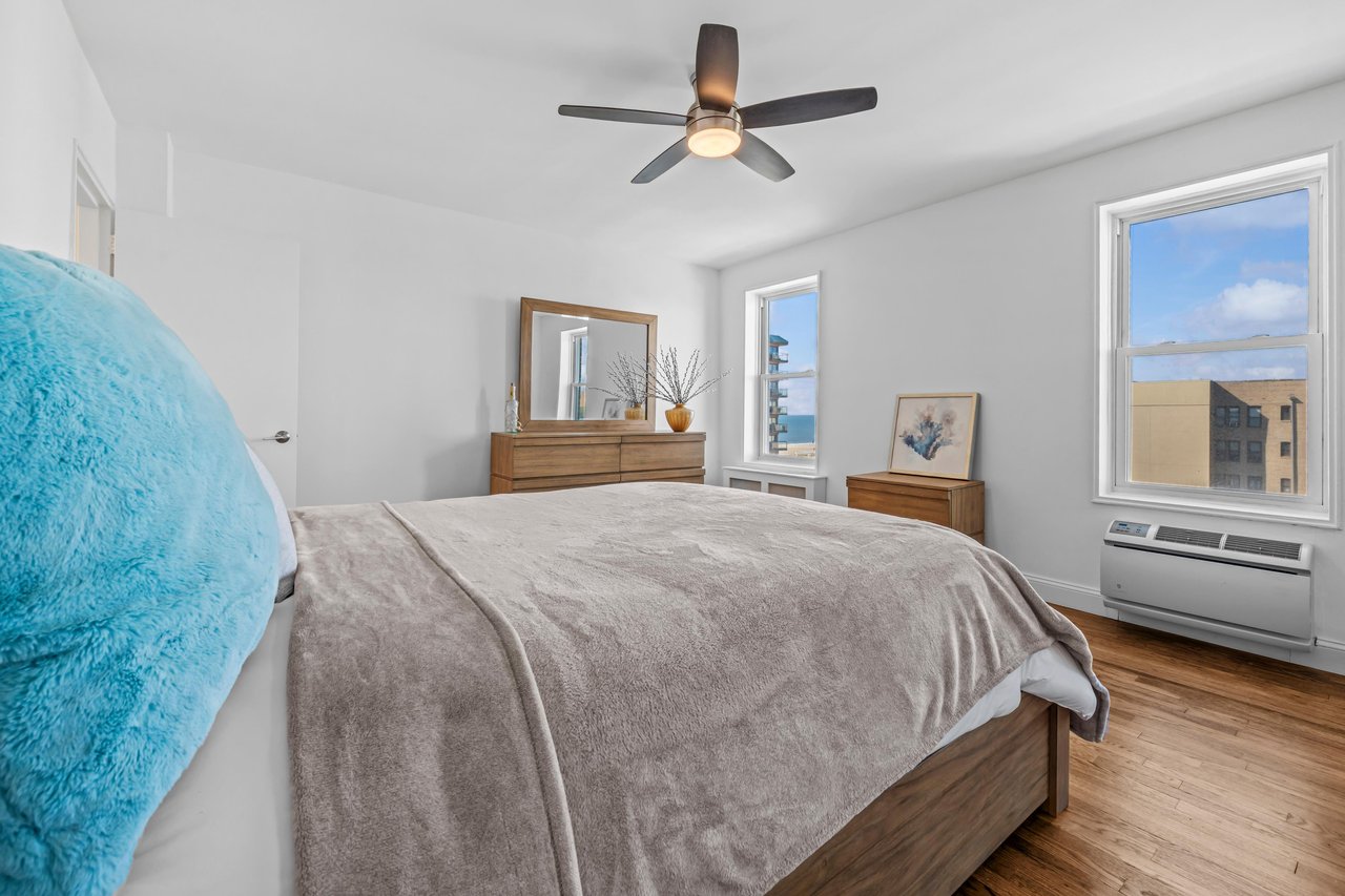 Bedroom with a gray-covered bed, blue fuzzy chair, wood furniture, and two bright windows.