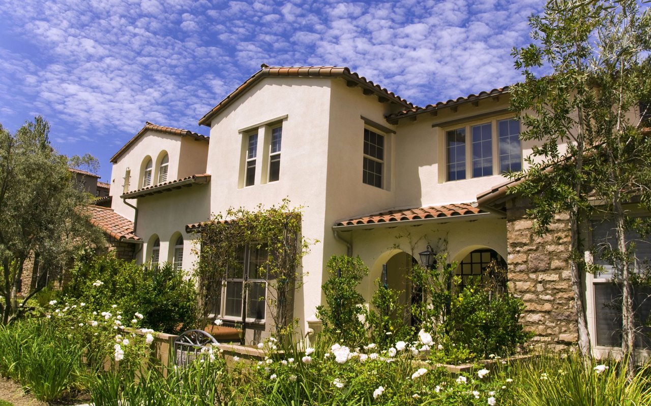 Two-story stucco house with a terracotta tile roof, arched entryways, and a flower-filled garden.