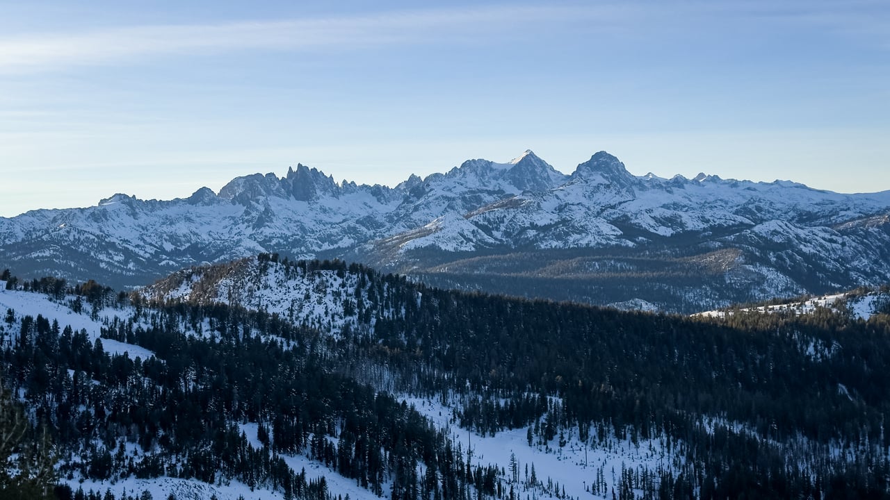 Snow-covered jagged mountains rise beyond forested slopes beneath a clear blue sky.