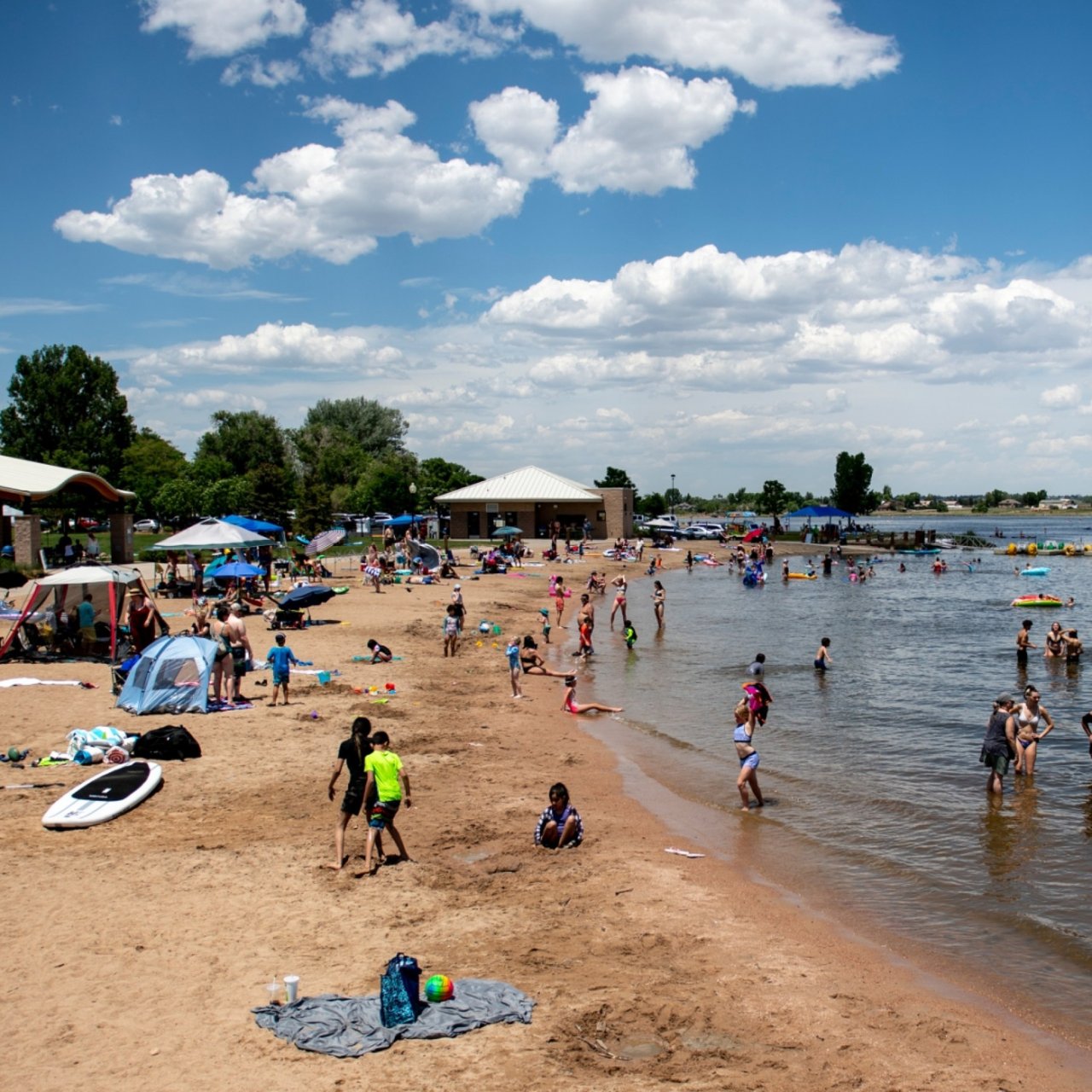 Crowded sandy beach beside a swimming area, with colorful umbrellas and a pavilion.