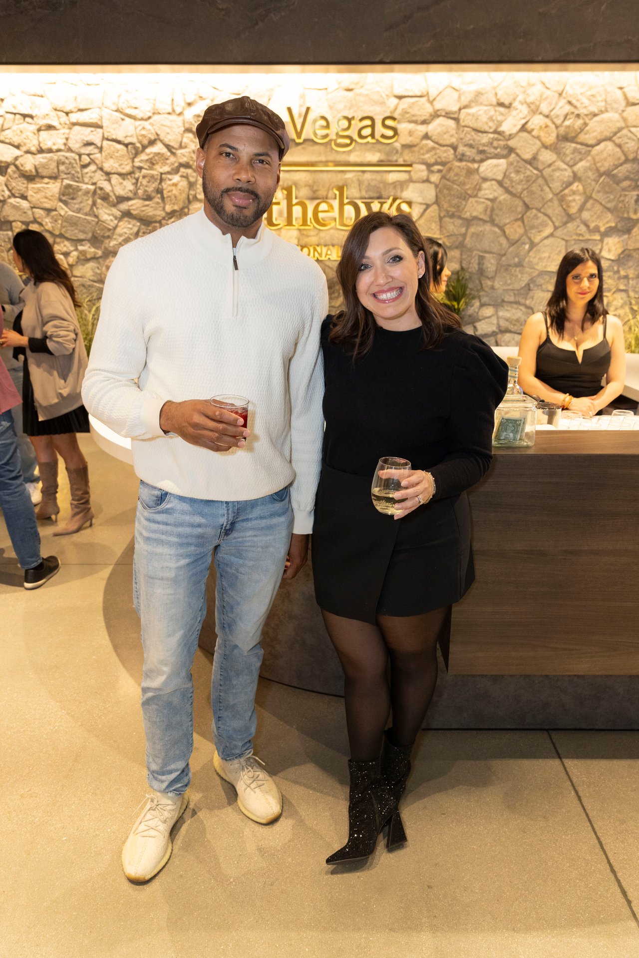 A man and woman pose with drinks at an event, with illuminated Vegas signage behind them.