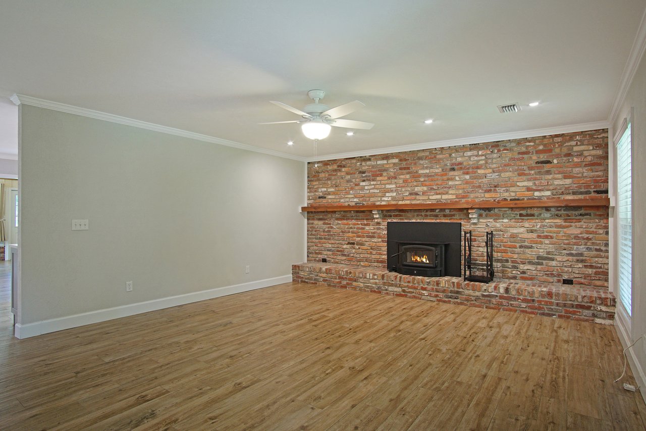 Spacious living room with wood floor and brick fireplace. A ceiling fan hangs above, and sunlight streams through windows, creating a cozy atmosphere.