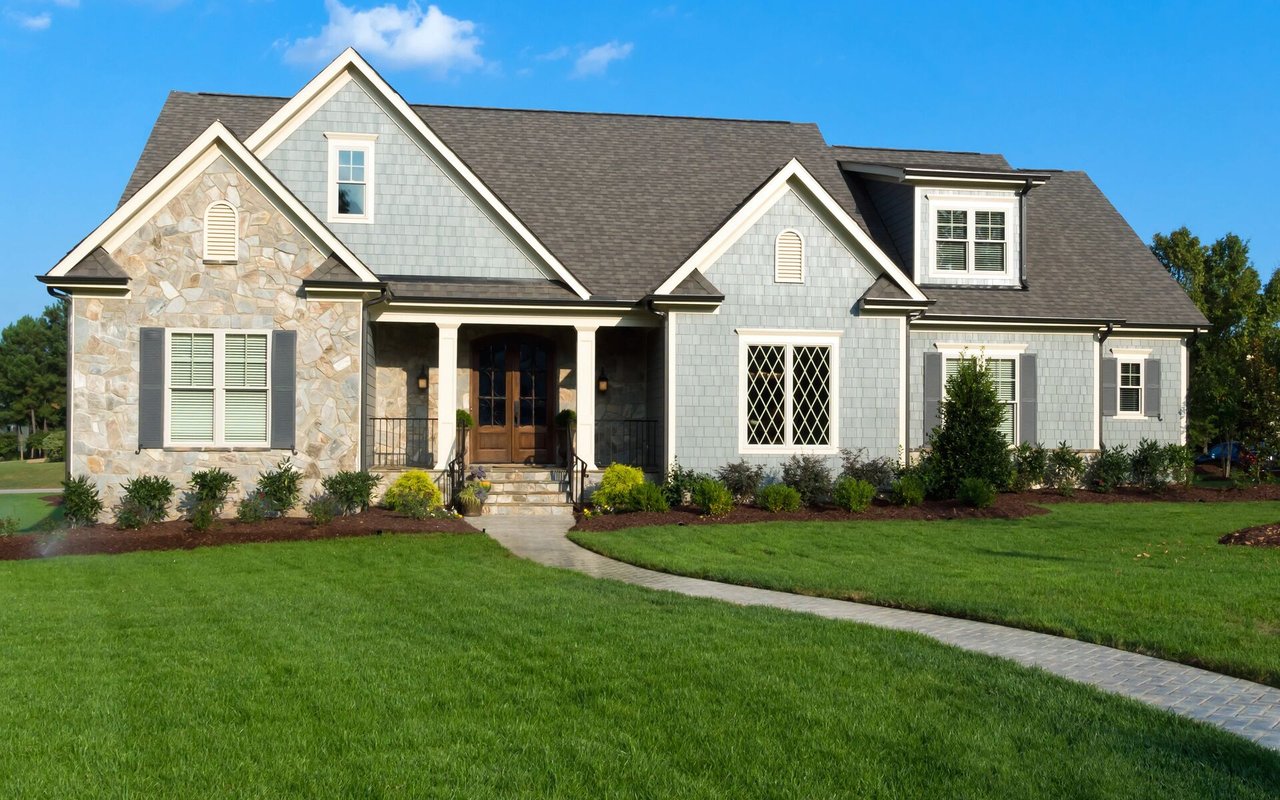 Gray-shingled house with stone accents, a covered porch, and a curved walkway across a broad lawn.