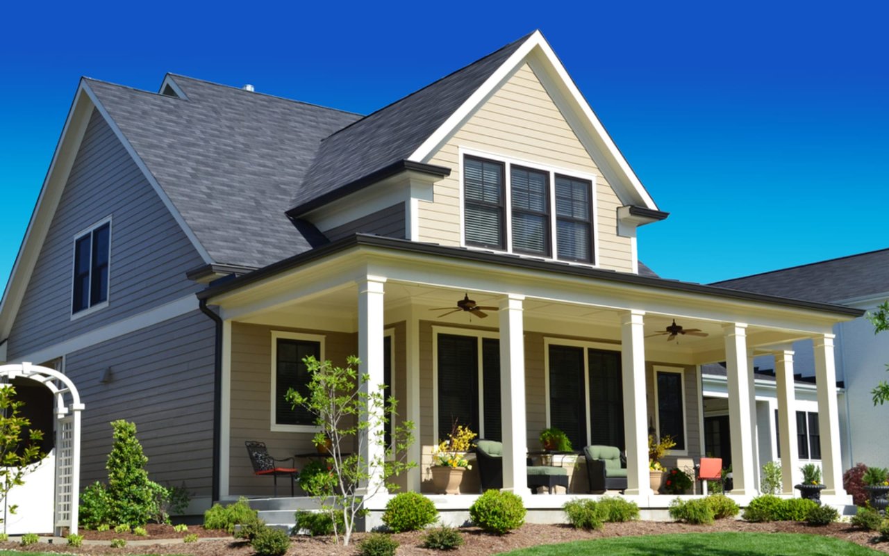 A beige colored Cape Cod style house with a large front porch and a blue sky in the background.