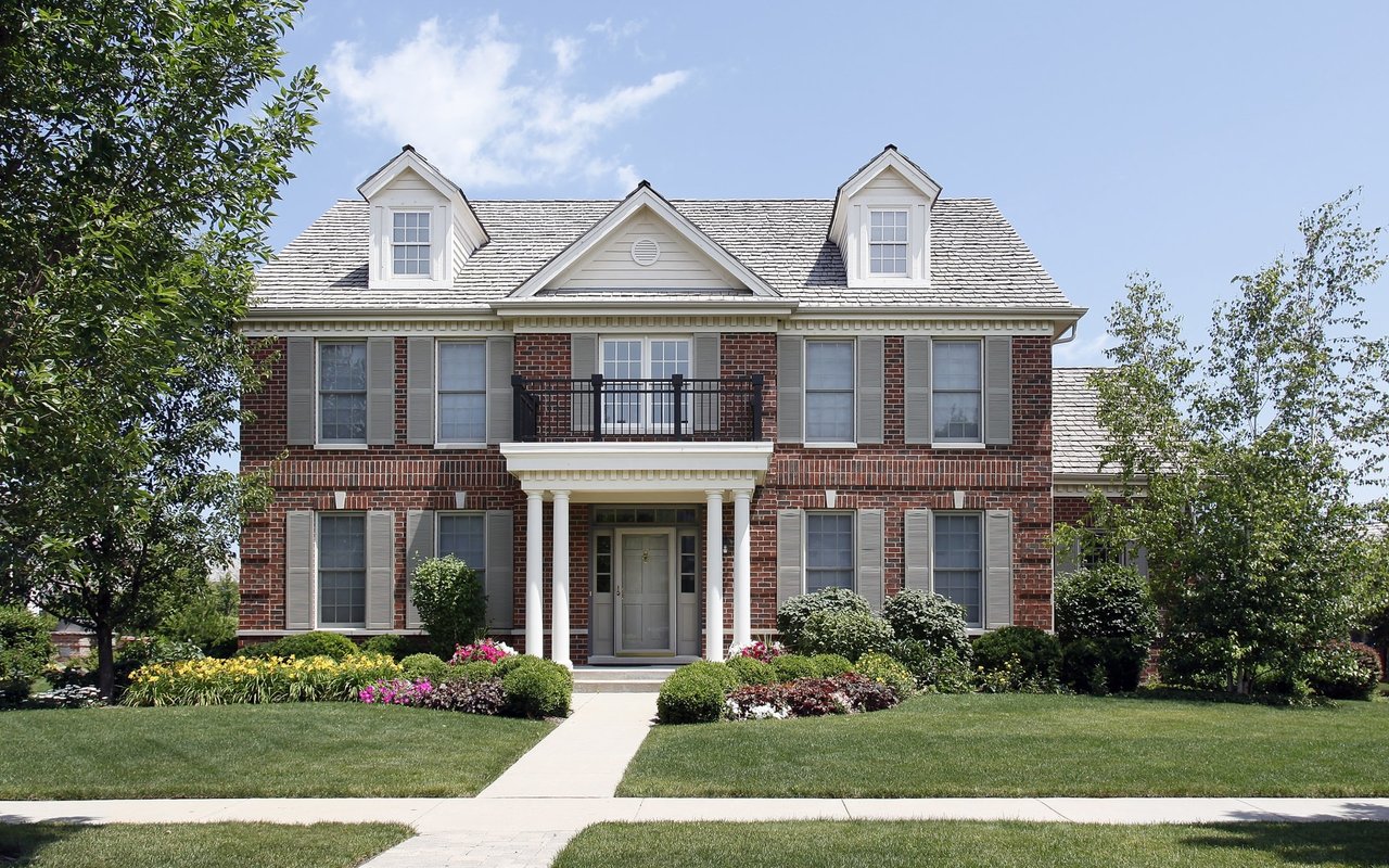 A two-story brick house with a balcony, columned entryway, covered porch, sidewalk, trees, and shrubs in the front yard.