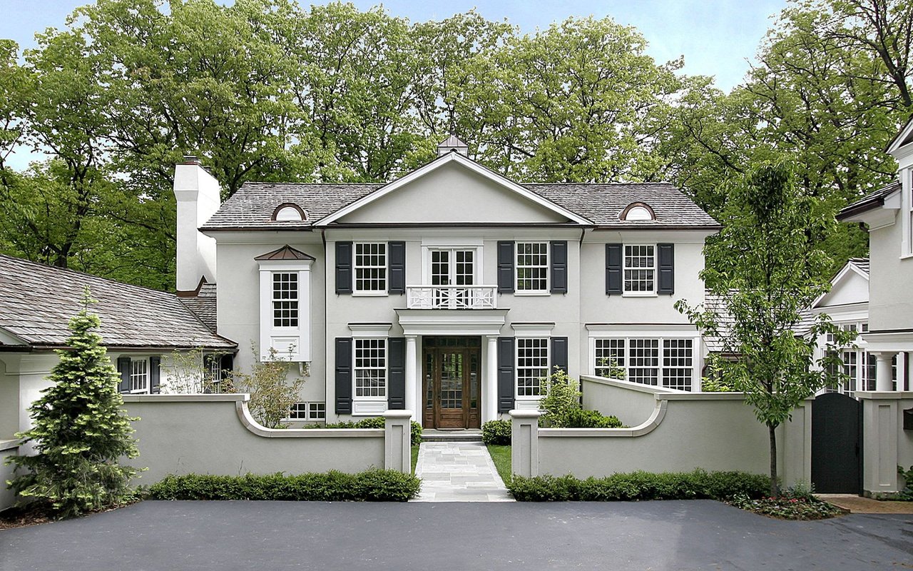 A large white house with black shutters sits on a tree-lined street