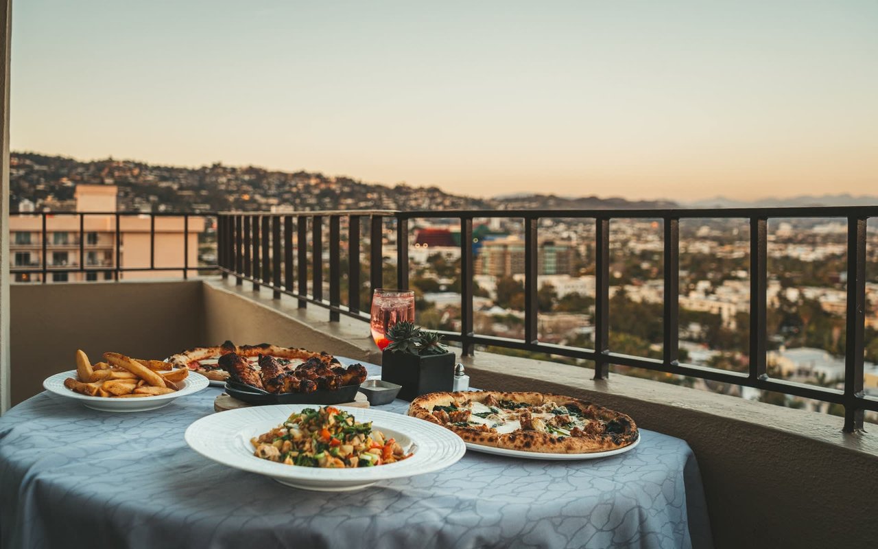  A table laden with a variety of food, including two pizzas, a bowl of pasta or salad, and a plate of french fries, is set on a balcony overlooking a sprawling cityscape at dusk.