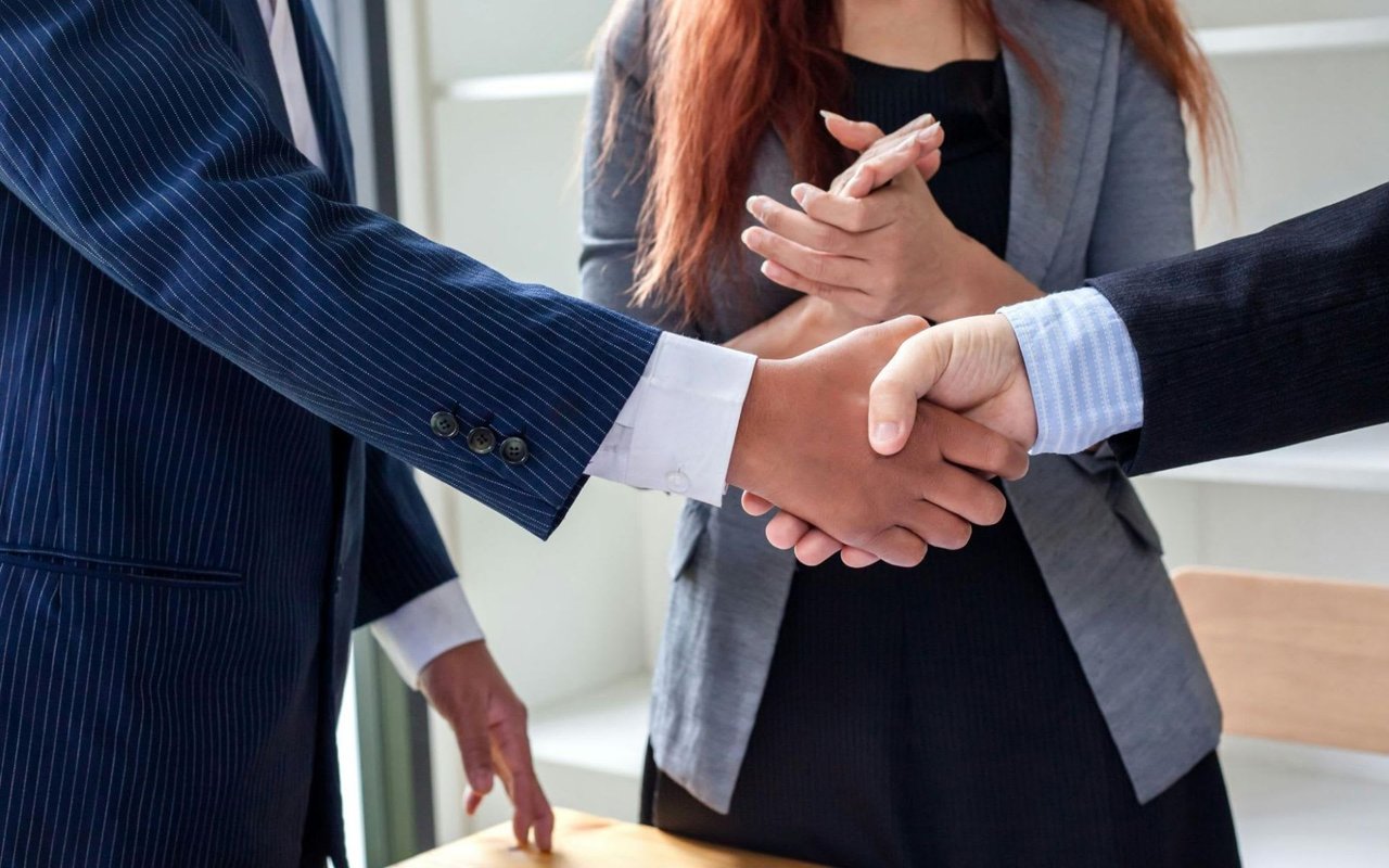Two men shaking hands with a woman clasping her hands in the background.