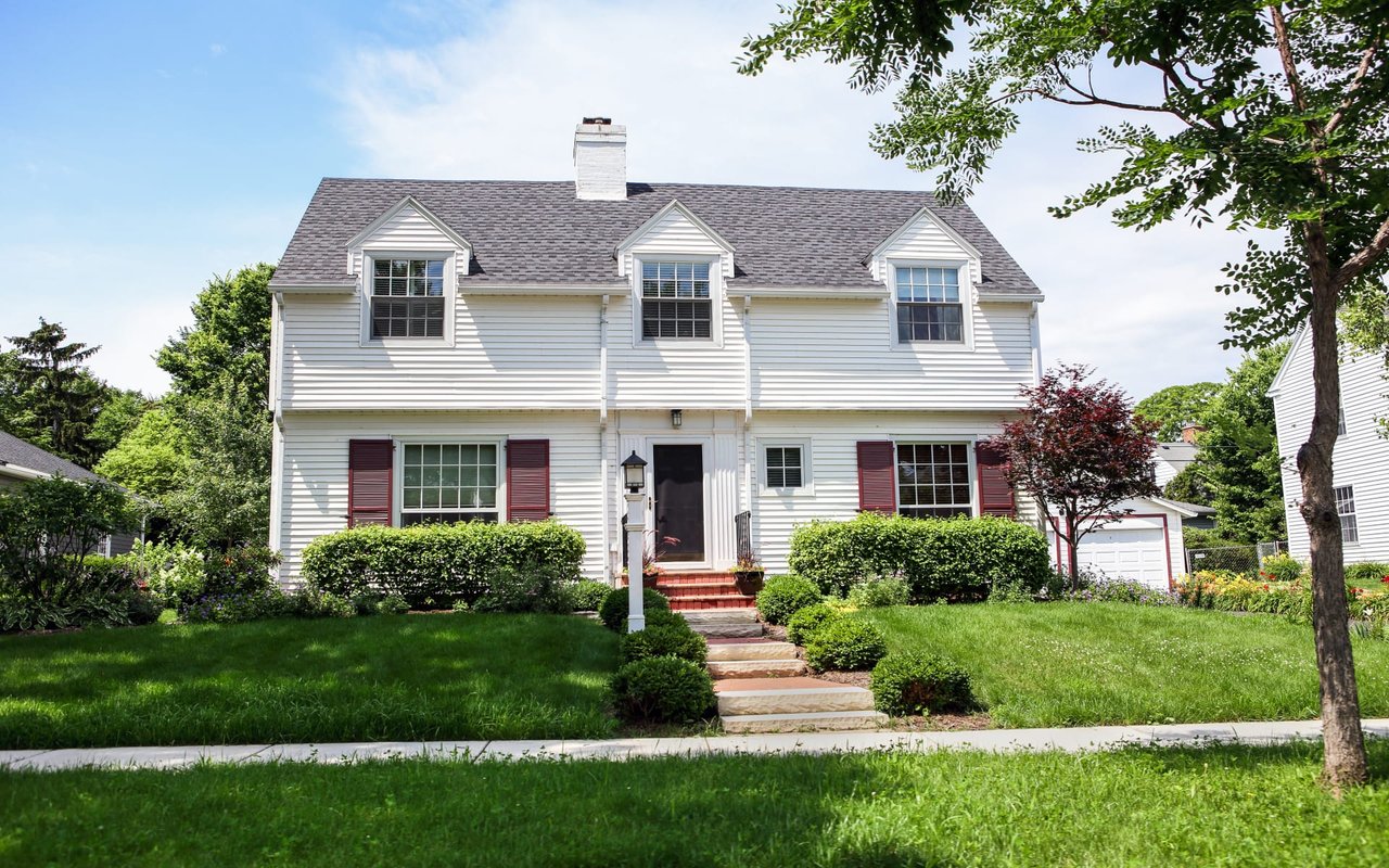 A two-story white house with red shutters, a brick stairway to the front door, and surrounded by green lawns and trees.