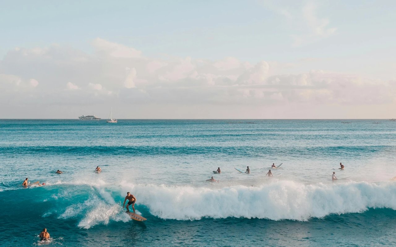Surfers ride a breaking wave offshore as swimmers gather in the turquoise water.